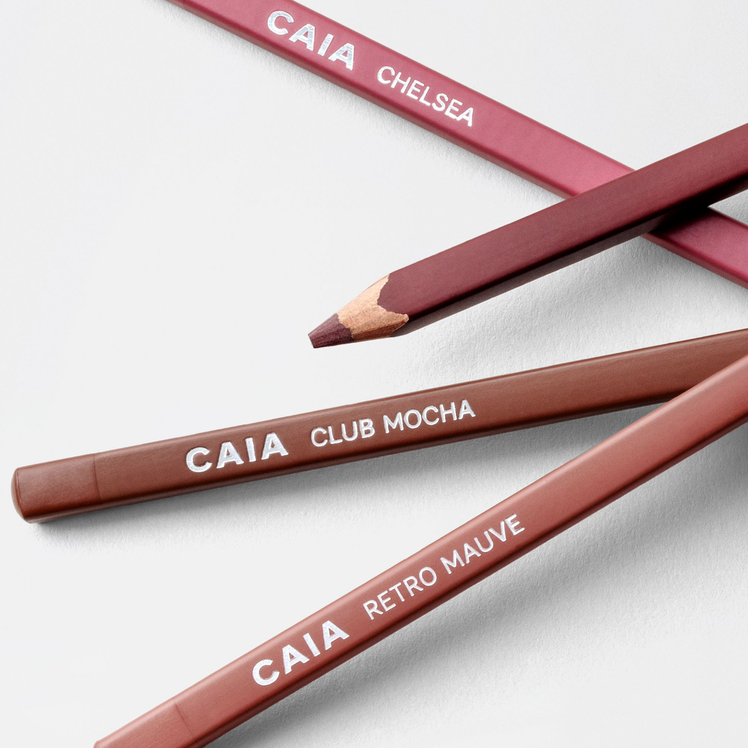 Close-up of four CAIA lip pencils in nude and pink shades, crossed on a clean background, with visible shade names including CLUB MOCHA and RETRO MAUVE.